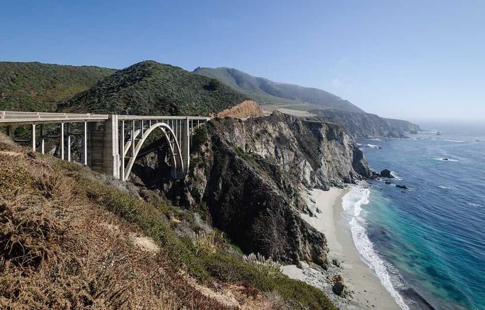 Concrete arch bridge over rugged coastal cliffs with sandy beach and blue ocean waves below.