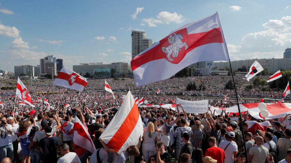 Large crowd with red and white flags gathered in a city square under a blue sky.
