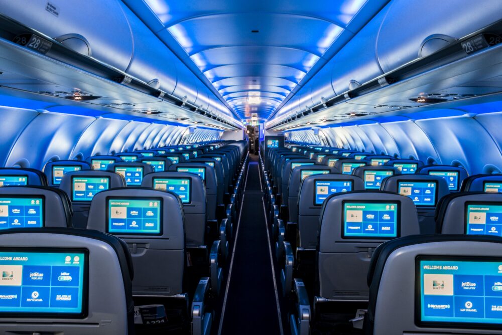 Rows of airplane seats with personal screens, illuminated in blue, viewed from the aisle toward the back.