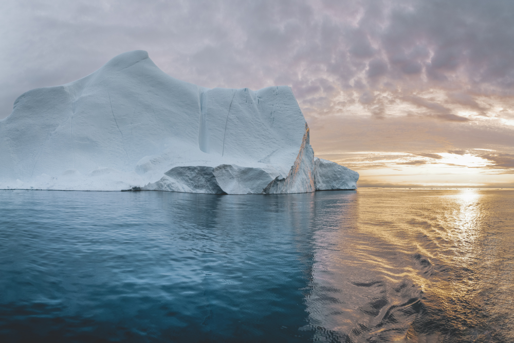 Large iceberg in calm ocean water with a colorful sunrise sky.