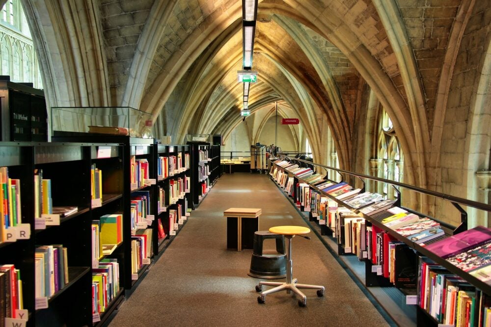 Library aisle with arched ceiling, black bookshelves filled with colorful books, and a stool in the foreground.