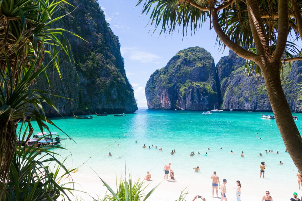 Tropical beach with people swimming, boats on turquoise water, and lush green cliffs in background.