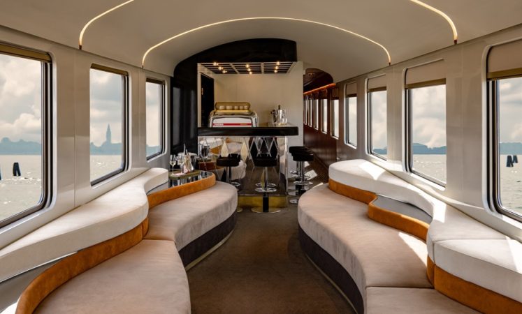 Luxurious train interior with curved seating, bar area, and large windows overlooking water and distant landmarks.