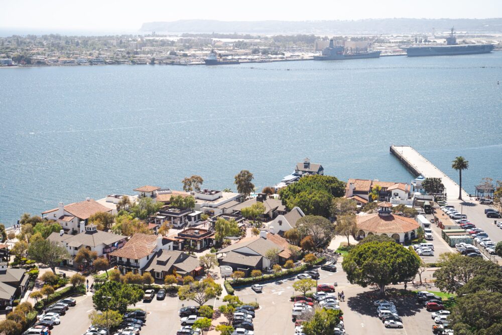 Aerial view of a coastal area with buildings, parked cars, and a pier extending into the water.