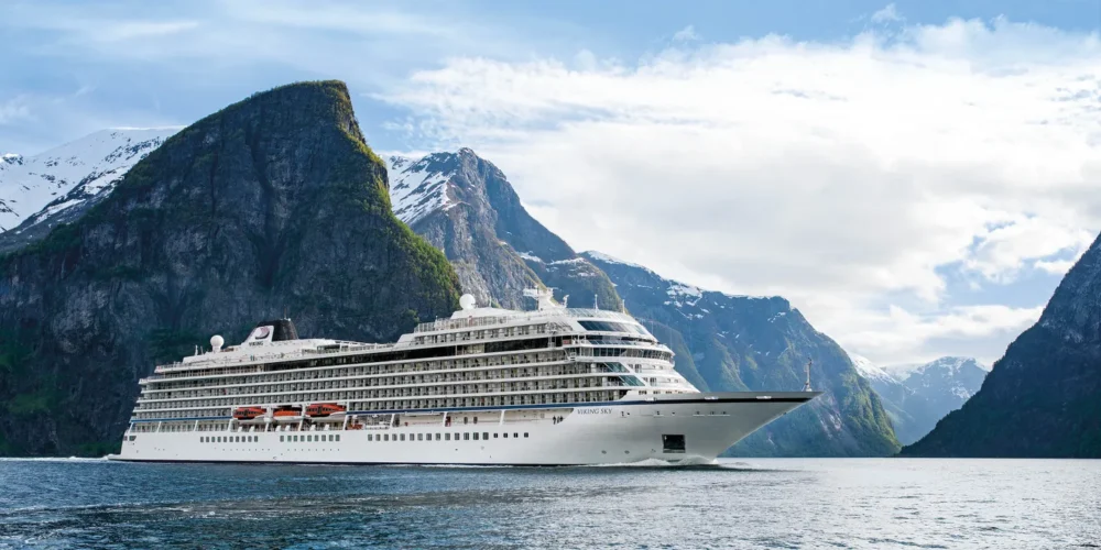 A cruise ship sailing through a fjord, surrounded by steep, snow-capped mountains under a partly cloudy sky.