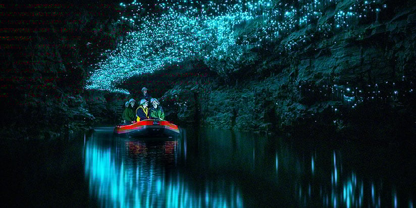 A boat with people floats through a dark cave illuminated by glowing blue lights on the ceiling.
