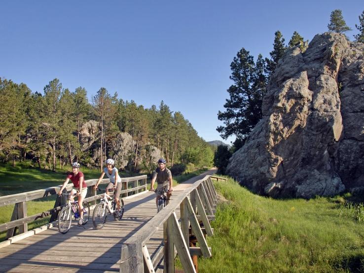 Three cyclists ride on a wooden bridge through a forested area with large rocks nearby.