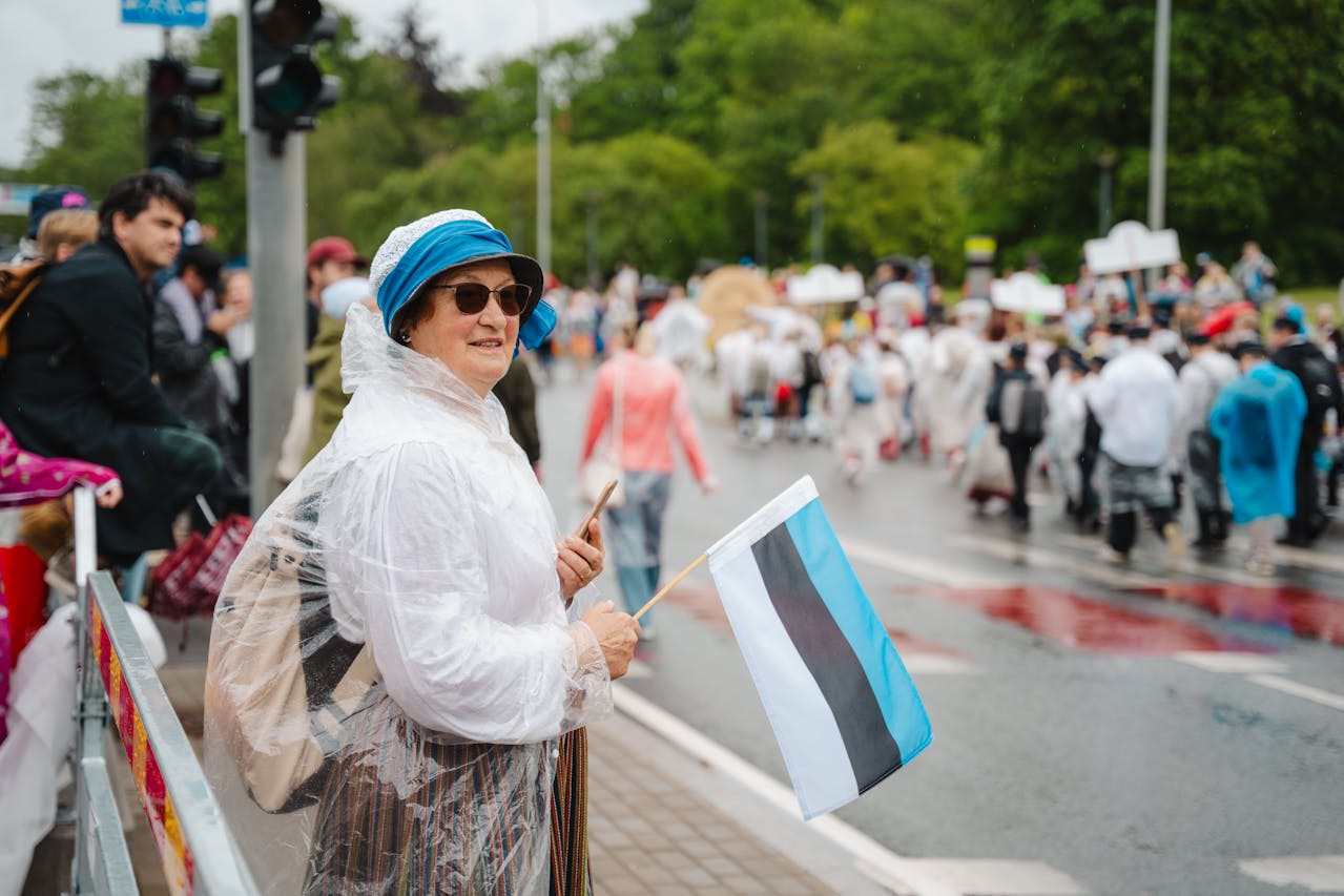 Woman in a blue hat and raincoat holding an Estonian flag at a street parade.
