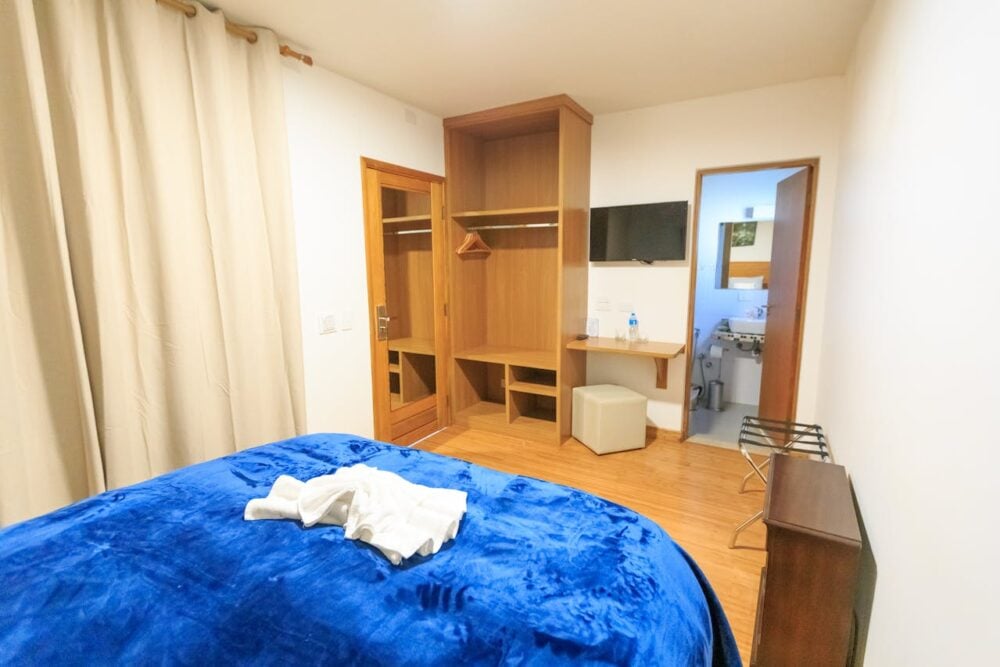 Bedroom with blue-covered bed, open closet, wall-mounted TV, small desk, and an open door to bathroom.