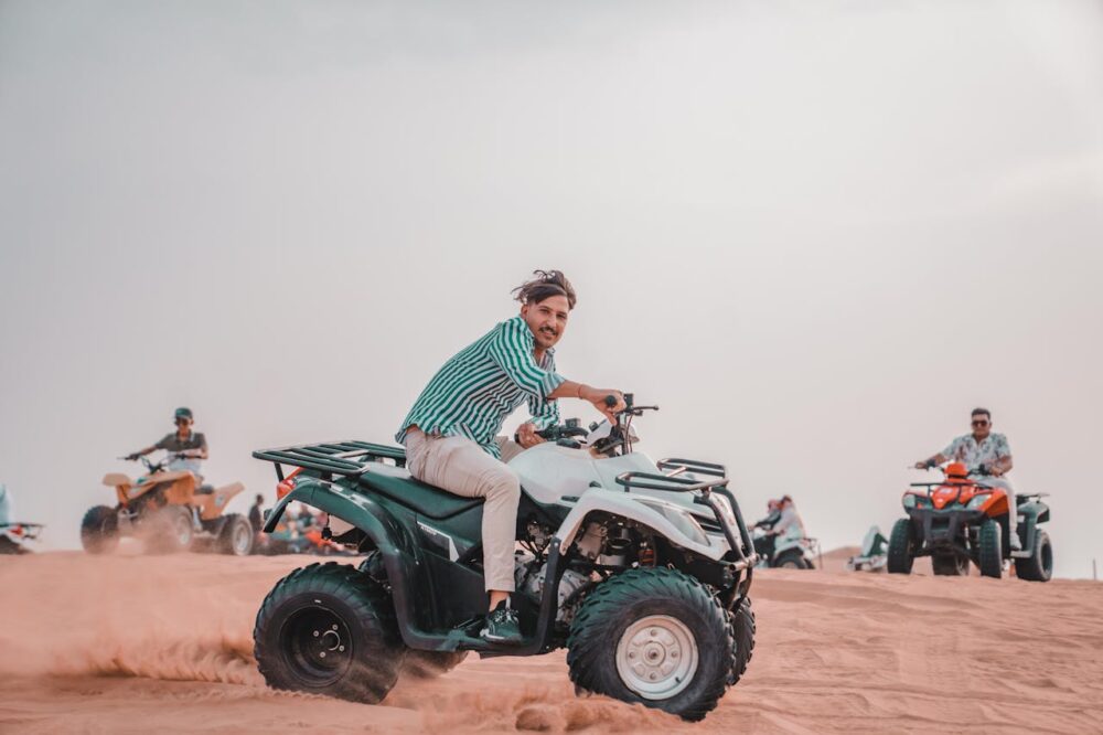 A person rides an ATV on sand dunes, with others on ATVs in the background.