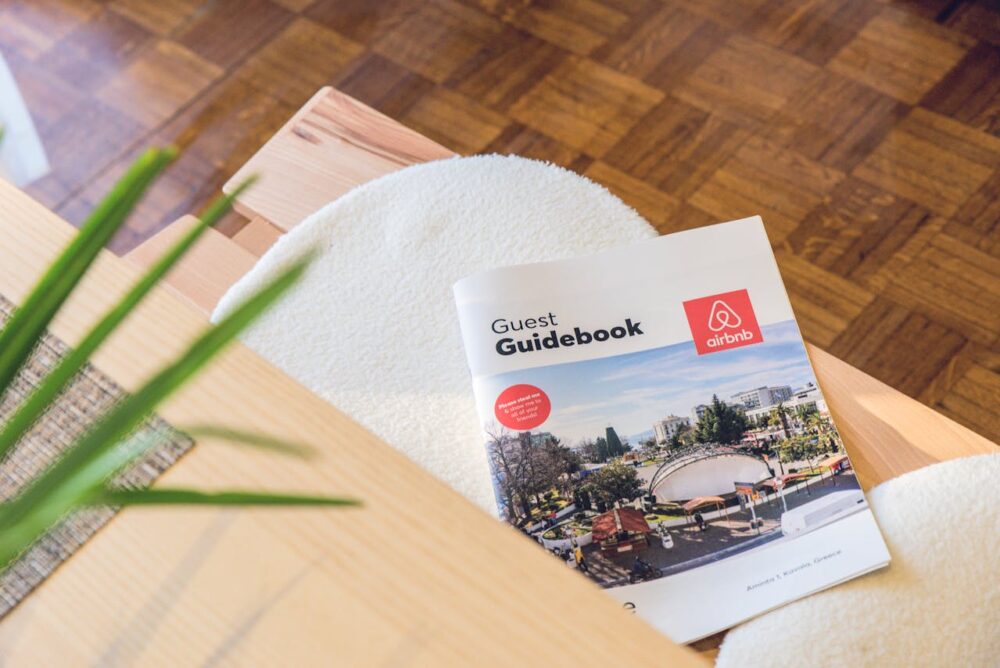 Airbnb guest guidebook on a wooden table with a round white cushion and plant nearby.
