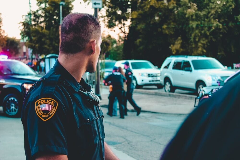 Police officer standing on a street, with two officers near parked cars in the background.