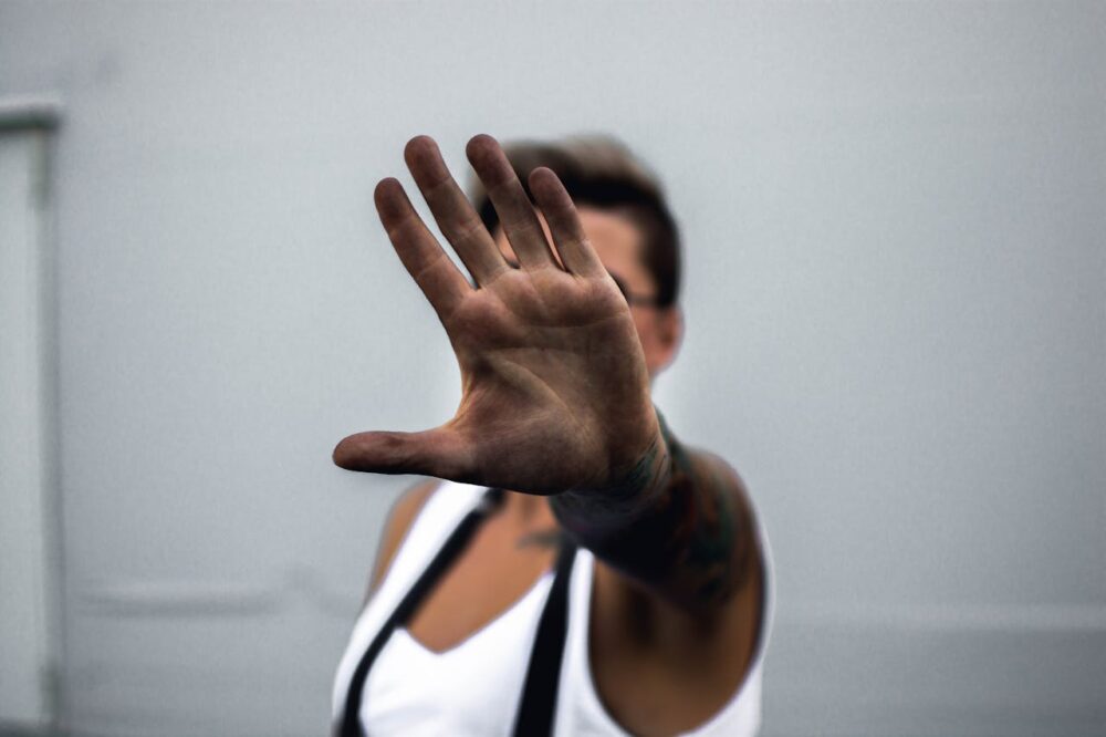 Person in white top with suspenders extending hand toward camera, palm facing forward, against a gray background.