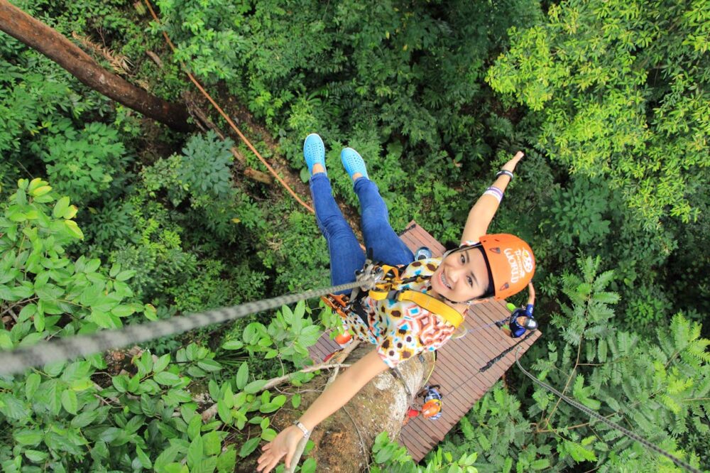A woman ziplining above a lush green forest wearing a safety helmet