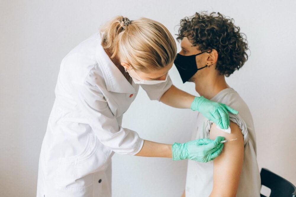 Health worker administering vaccine to seated person wearing a mask; both wearing protective attire.