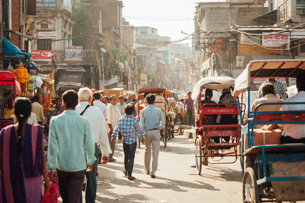Busy street scene with people walking, rickshaws, and shops displaying colorful signs and wares.