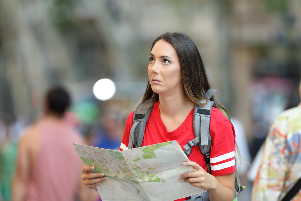 A woman with a backpack in a crowded street holds a map, looking upwards.