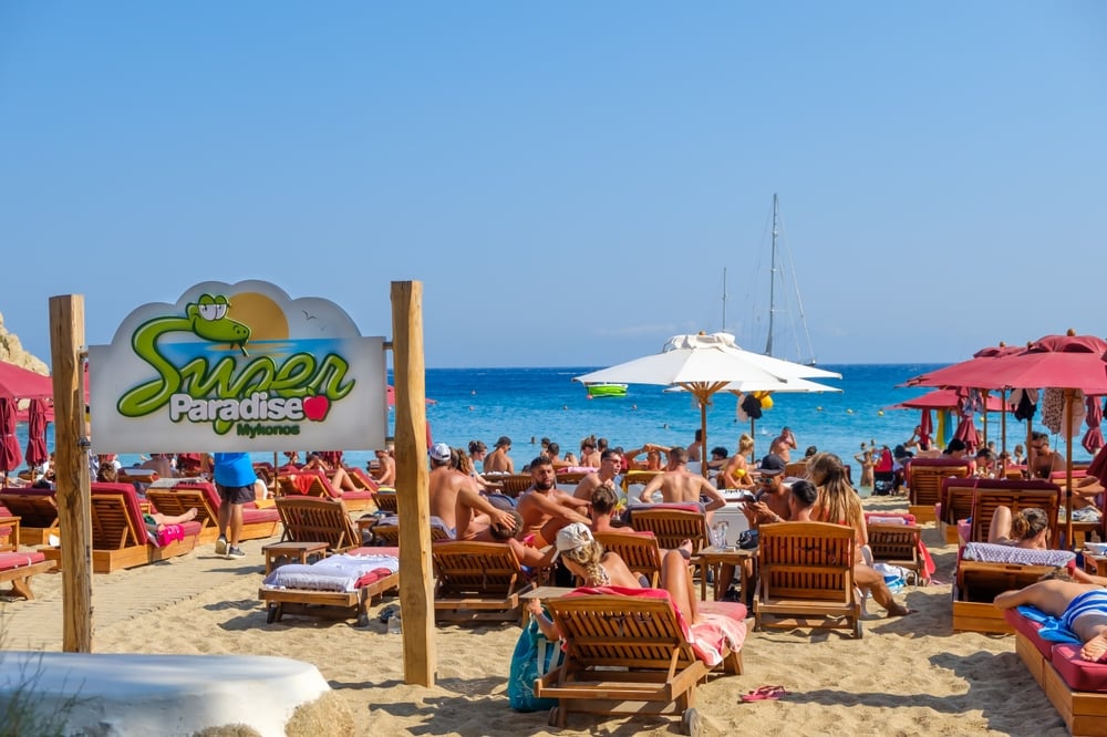 People relaxing on sun loungers under umbrellas at a crowded beach with a "Super Paradise" sign.
