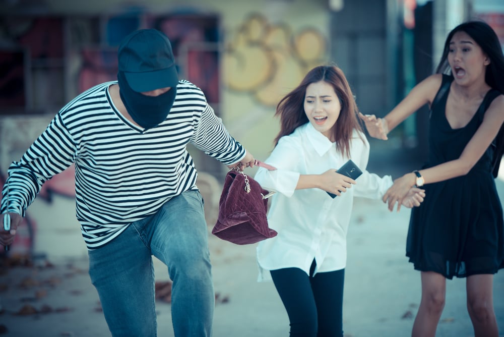 A masked person running, holding a woman's purse while two women react in surprise.
